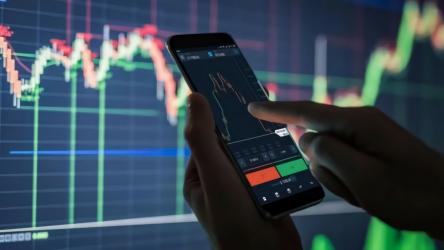 Image of a data graph and a mobile phone in someone's hands, which displays financial information.