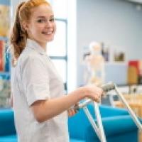 A female student smiling at the camera as she carries a walking frame to a patient