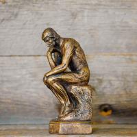 A small replica of 'The Thinker' - a bronze sculpture by Auguste Rodin depicting a man hunched over on a rock in contemplation - set against a backdrop of wood panels.