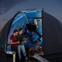 A small, domed rooftop observatory opens onto a starry night sky. Inside the observatory, two students use a telescope and a laptop to collect data.