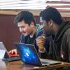 2 students at a cafe table chatting with laptops.