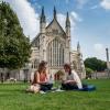 Two students sit talking on the grass outside a large, ornate cathedral.